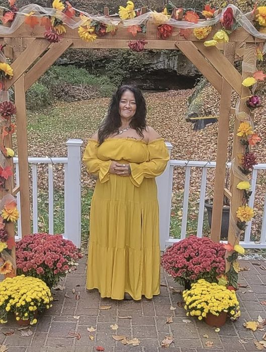 A reviewer stands gracefully in a flowing, off-the-shoulder dress beneath a flower-decked wooden arch, positioned on a brick path surrounded by potted chrysanthemums.