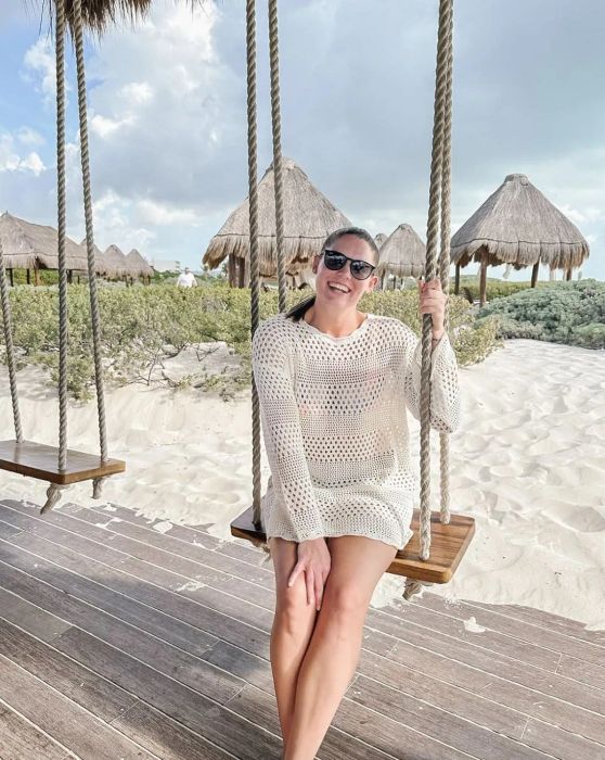 A person in a knit dress and sunglasses sits on a swing at a beach resort, smiling