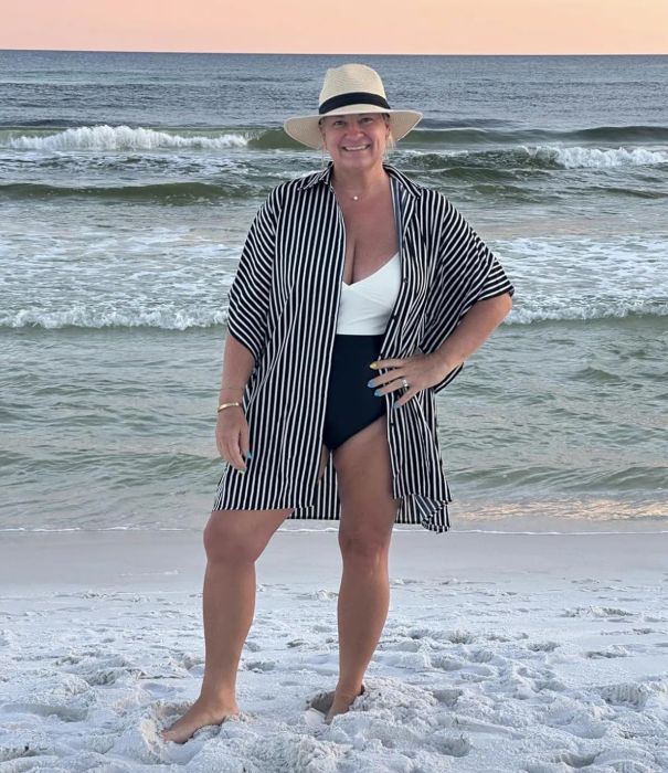A reviewer in a swimsuit and striped cover-up, wearing a hat, stands barefoot on a beach with waves crashing in the background