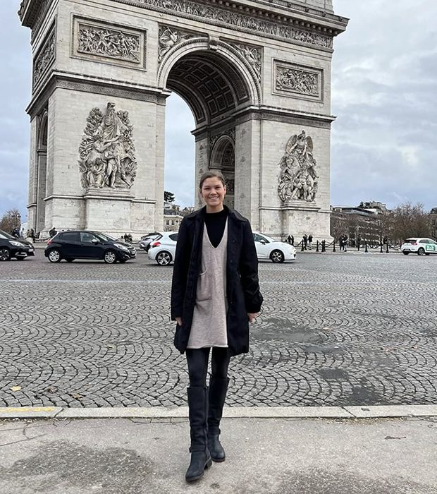 A reviewer stands in front of the Arc de Triomphe in Paris, dressed in a long coat over a dress with leggings and boots, beaming at the camera.