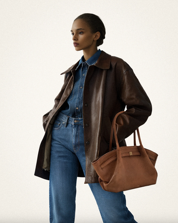 Model in a denim shirt, jeans, and a dark leather coat, carrying a large brown tote bag during a fashion shoot