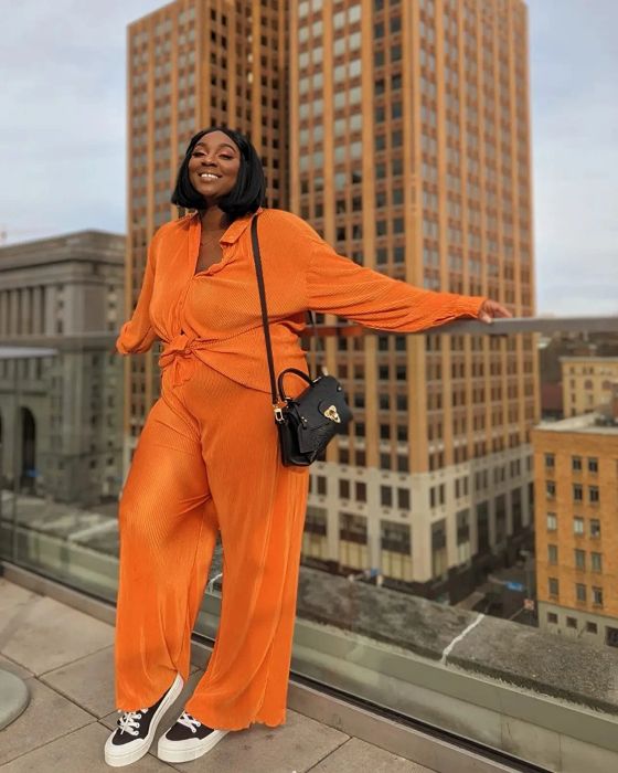 A reviewer in a fashionable orange jumpsuit, carrying a shoulder bag, strikes a confident pose in an urban setting