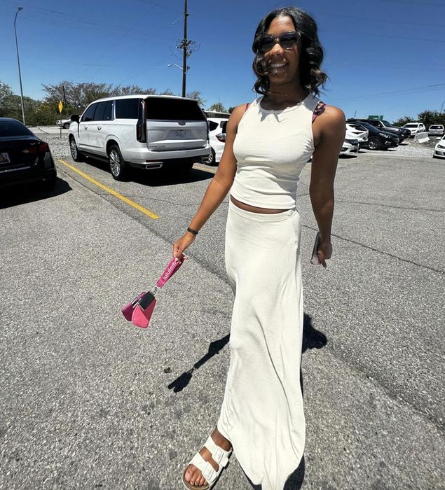 A reviewer is outdoors in a parking lot, sporting a sleeveless, long white dress, sandals, and sunglasses, while holding a pink bag.