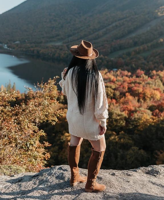 A reviewer with long hair, wearing a hat, sweater, and knee-high boots, stands on a rock, gazing over a picturesque landscape with a lake and mountains in the background