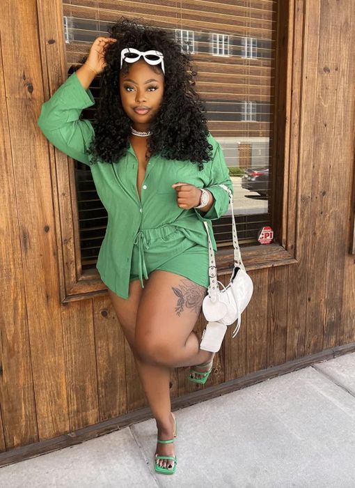 A curly-haired woman wearing glasses, a green shirt, shorts, sandals, and a white bag poses against a wood-paneled wall