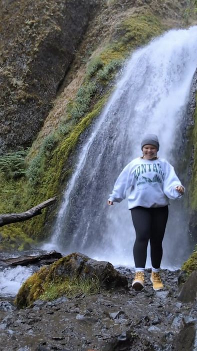 A person smiling and walking by a waterfall, dressed in a sweatshirt, leggings, boots, and a beanie.