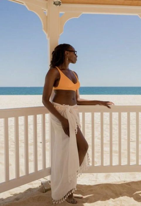 A reviewer stands under a gazebo near a sandy beach, wearing sunglasses, an orange bikini top, and a white sarong