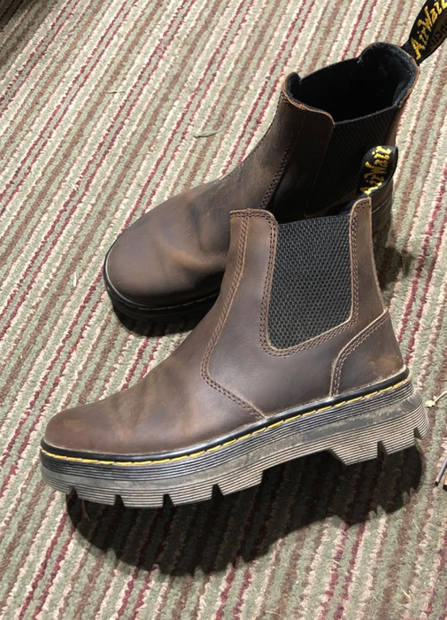 A pair of brown Dr. Martens Chelsea boots featuring thick, textured soles, displayed on a striped surface.