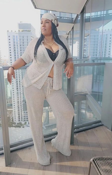 A reviewer poses on a balcony wearing a crochet cardigan, spaghetti strap top, and pants, with skyscrapers visible in the background