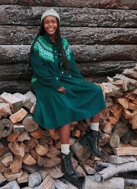 A smiling person sitting on stacked logs, dressed in a warm knit sweater, a long skirt, boots, and a beanie