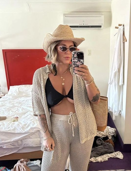 A reviewer in a crochet outfit with a bikini top underneath, wearing a straw hat and sunglasses, takes a mirror selfie in a cluttered room