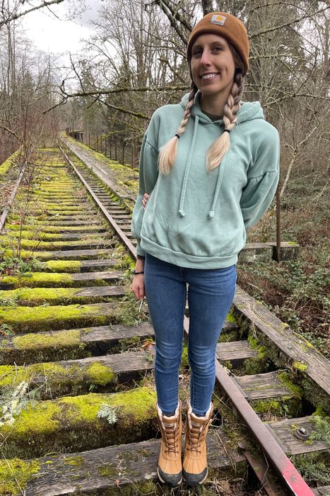 A reviewer with braids, dressed in a casual hoodie, jeans, and brown boots, standing on an old, moss-covered railway track surrounded by a forest.