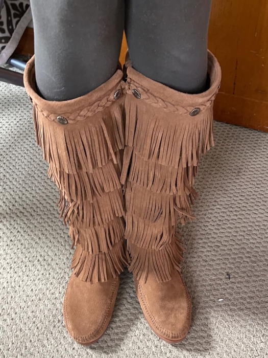 A close-up of a person’s legs, featuring knee-high suede moccasin boots with fringe details.