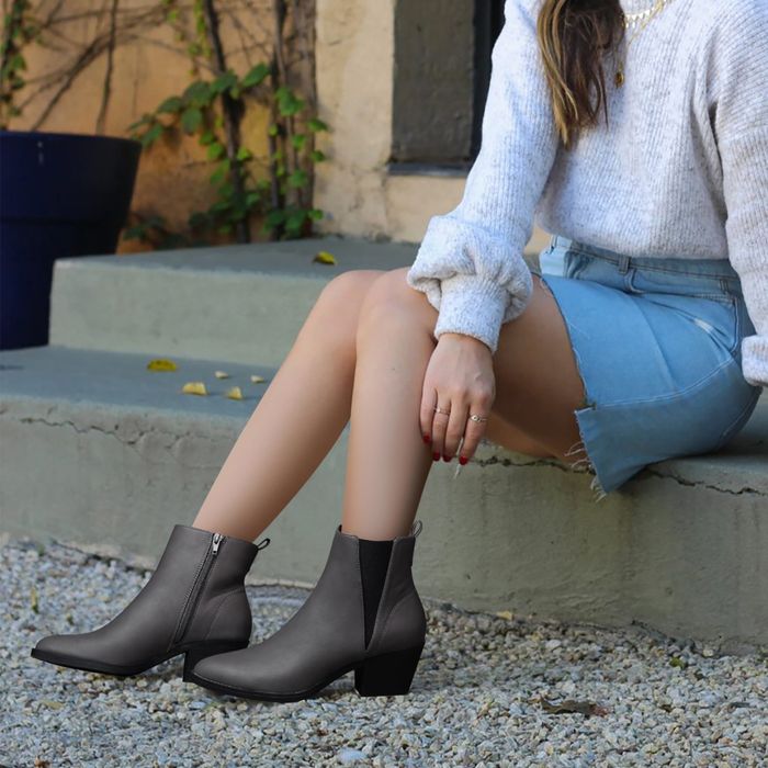 A person sitting on a ledge, wearing a gray sweater, denim skirt, and ankle boots, showcasing a casual fall look