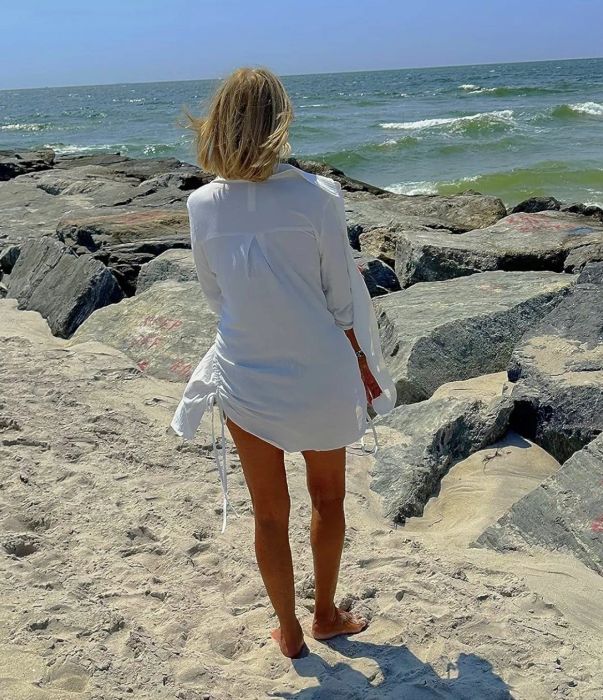 A person in a breezy dress stands barefoot on a sandy beach, facing the ocean with rocks scattered along the shoreline