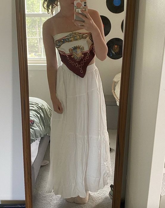 A person taking a mirror selfie in a bedroom, wearing a strapless top and a long white skirt.