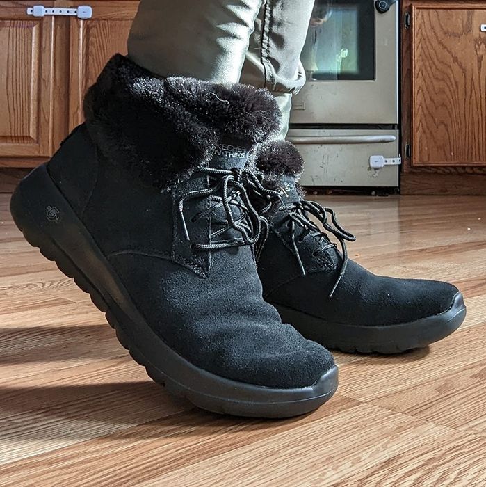 A reviewer wearing black winter boots with fur lining, standing on a hardwood floor in a kitchen setting.