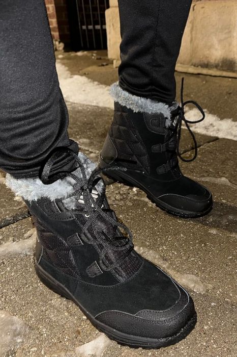 A reviewer wearing black winter boots with fur trim, standing on a snowy sidewalk.
