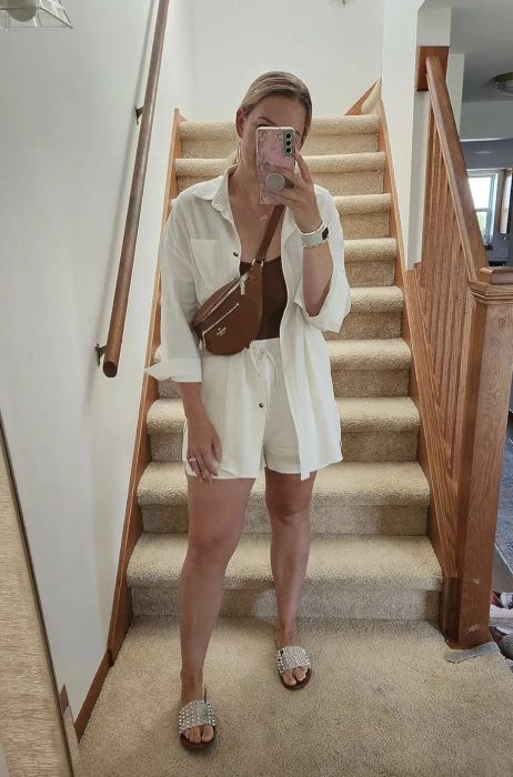 A woman stands on stairs, taking a mirror selfie. She’s wearing a white open shirt, a brown top, white shorts, and sandals, carrying a brown shoulder bag with a pink phone case