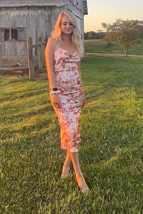 A reviewer wearing a floral dress stands on the grass near a barn during sunset.