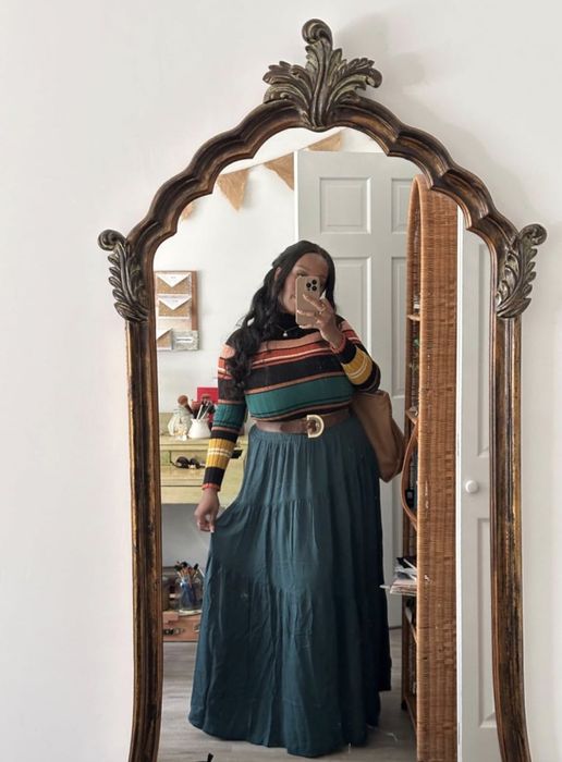 A person takes a mirror selfie in a home setting, wearing a striped sweater and a long skirt.