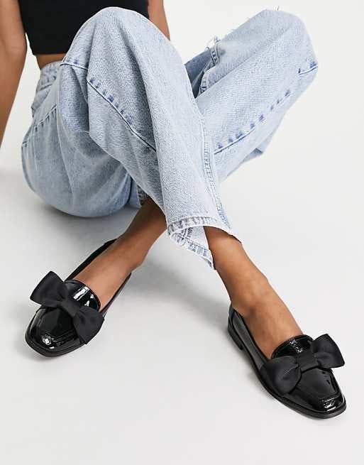 Person styling light-wash jeans with black patent leather loafers featuring large bows