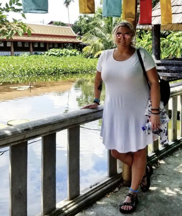 A woman in a white dress, sunglasses, and sandals stands by a railing overlooking a pond surrounded by greenery and colorful flags