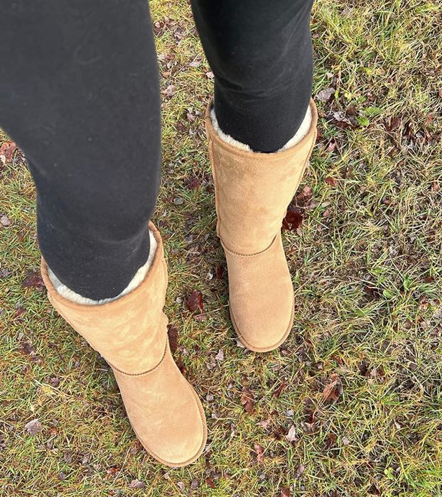 A pair of tan suede boots and black leggings standing on a grassy field scattered with fallen leaves.