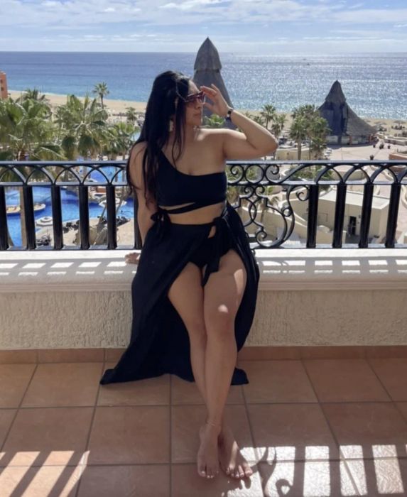 A reviewer in a sleeveless black outfit and sunglasses sits on a balcony, enjoying a sunny seaside view in the background