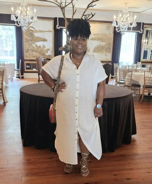 A person in a white button-down dress and strappy sandals stands in a luxurious room adorned with chandeliers and decorative wall art