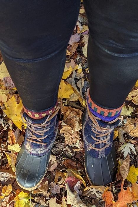 A reviewer wearing plaid boots and leggings, standing on a bed of colorful autumn leaves scattered on the ground.