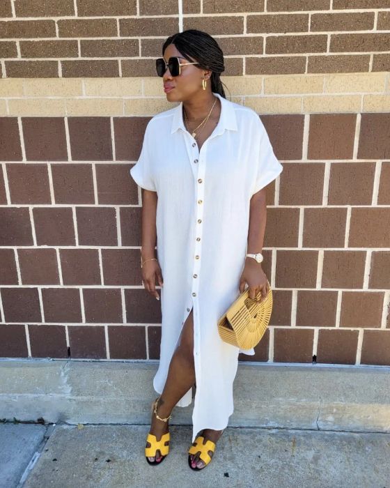A reviewer in a white buttoned dress and yellow sandals, wearing sunglasses, poses by a brick wall while holding a fan-shaped handbag