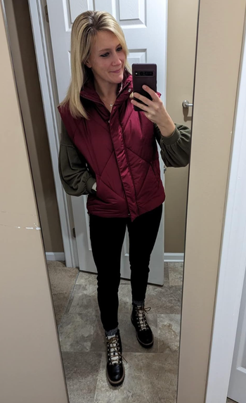 A model takes a mirror selfie wearing a puffer vest over a sweater, black pants, and boots, standing in a modern bathroom.