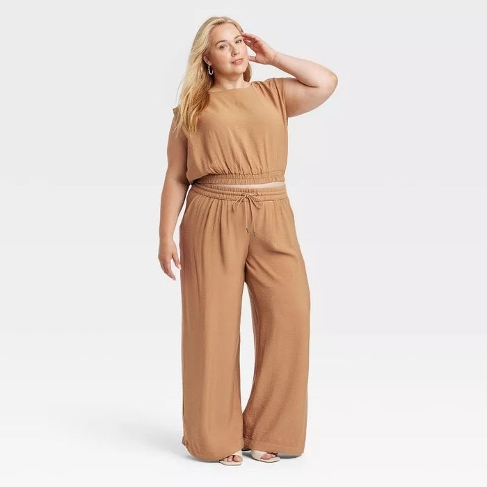 A model posing in a loose-fitting, short-sleeved jumpsuit featuring a tied waistband, standing casually