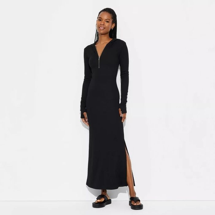 A person standing against a plain background, wearing a long-sleeve, ankle-length black zip-up dress with a slit and black open-toe sandals
