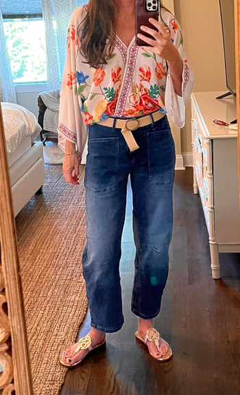 A person captures a mirror selfie while wearing a floral blouse, blue jeans, and sandals with intricate cut-out details.