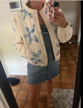 A person in a quilted jacket featuring a floral design, layered over a denim dress, taking a mirror selfie in a shopping article.