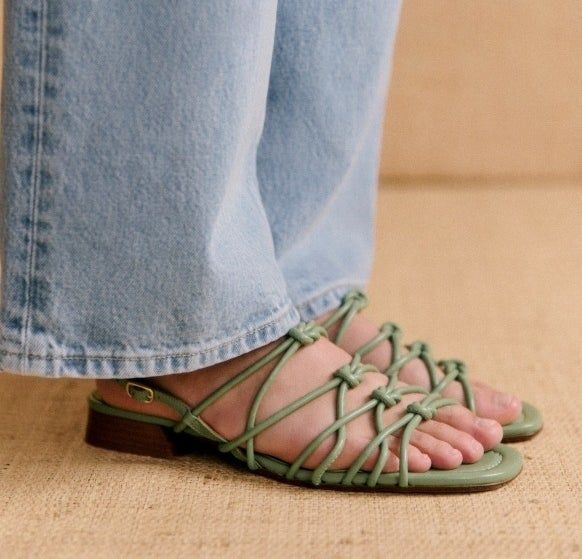 A close-up of someone's feet, dressed in light blue jeans and stylish green strappy sandals with low wooden heels.