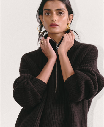A model wearing a ribbed, oversized black sweater with a half-zipper, accessorized with large hoop earrings and minimal makeup, her hair styled in a loose updo.