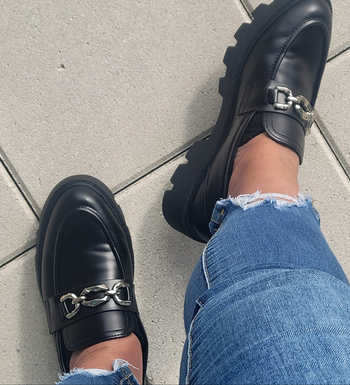 A person wearing black loafers with chain accents, paired with ripped blue jeans.