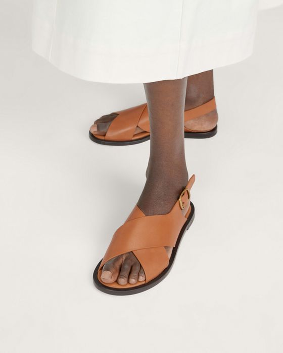 This model is wearing tan leather sandals with crisscross straps and a buckle at the back.
