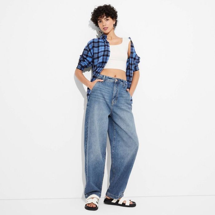 A person wearing a blue plaid shirt, a white crop top, baggy jeans, and black-and-white sandals
