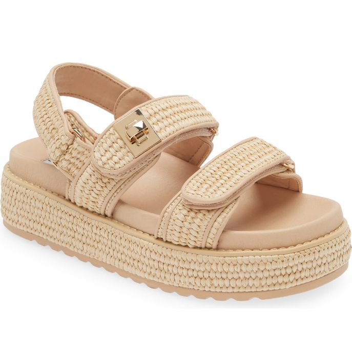 Close-up image of a beige woven platform sandal featuring a thick sole and adjustable straps. The sandal includes a textured design and a metallic buckle detail.