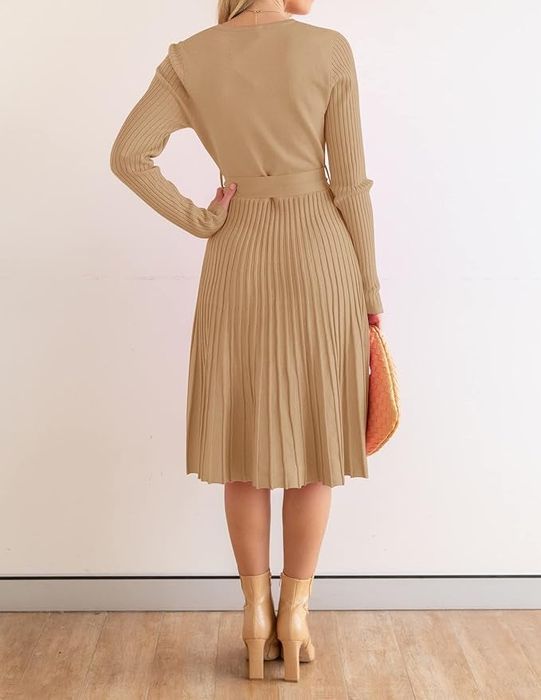 A back view of a person wearing a pleated knee-length dress, holding a woven handbag, and standing indoors in front of a white wall.