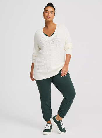 A person in a relaxed pose wearing an oversized white sweater, green joggers, and matching green sneakers.