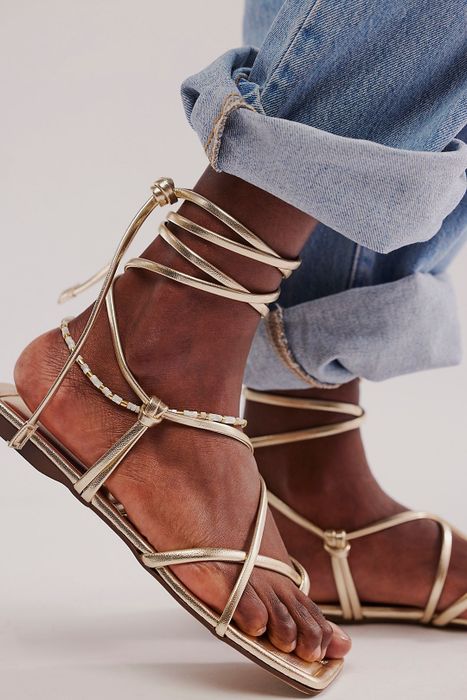 Close-up image showing a person's feet in gold wrap-around sandals paired with rolled-up jeans.