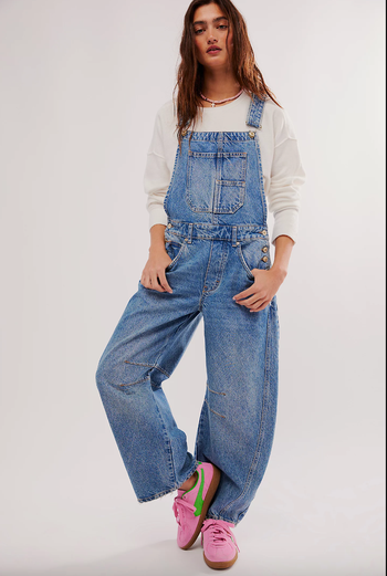 A person wearing denim overalls, a white long-sleeve shirt, and pink sneakers, confidently posing.