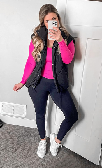 A model taking a mirror selfie in a sporty casual outfit: a black puffer vest, pink top, black leggings, and white sneakers.