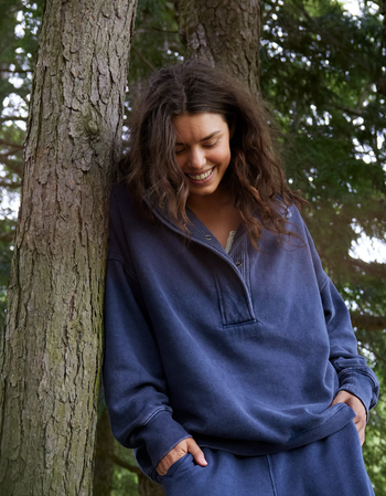 A person smiles while leaning against a tree, wearing an oversized hoodie and relaxed pants for a casual look.