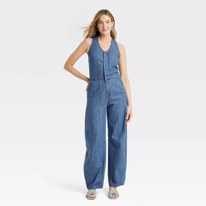 a model wearing a sleeveless denim jumpsuit and sandals, smiling confidently at the camera
